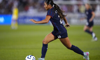 A woman in a blue jersey dribbles the ball on a soccer field